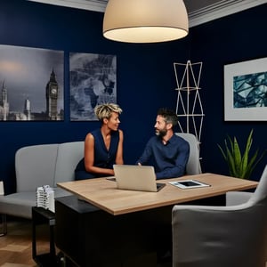 Zwei Geschäftsleute besprechen Projekt am Laptop, im Hintergrund moderne Büroeinrichtung mit London-Skyline-Bild