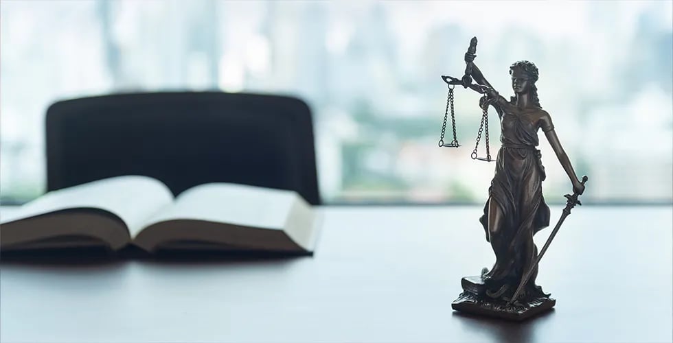 Auf einem Tisch liegt ein Gesetzesbuch, daneben steht eine kleine Statue der Justitia.
