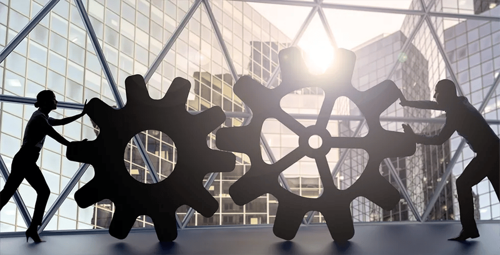 In front of a window, you see the silhouettes of two people connecting large gears.