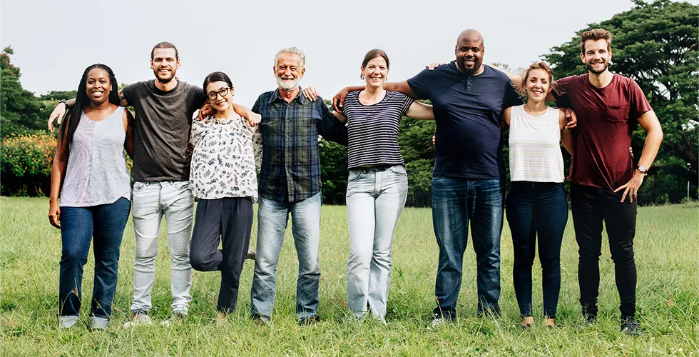 Eight people from different cultures and with different nationalities stand arm in arm on a meadow.