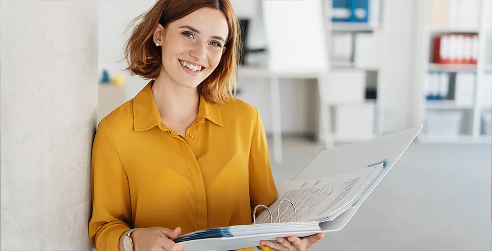 A young woman holds a folder with legal translations in her hand and smiles at the camera.