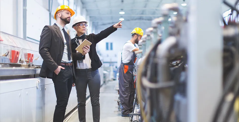 Two people are standing in front of a large machine in the production hall. One is holding a tablet with a mechanical engineering translation.