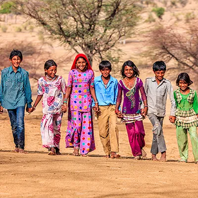 Sieben Kinder in bunten Gewändern gehen Hände haltend und lachend über einen Sandplatz.